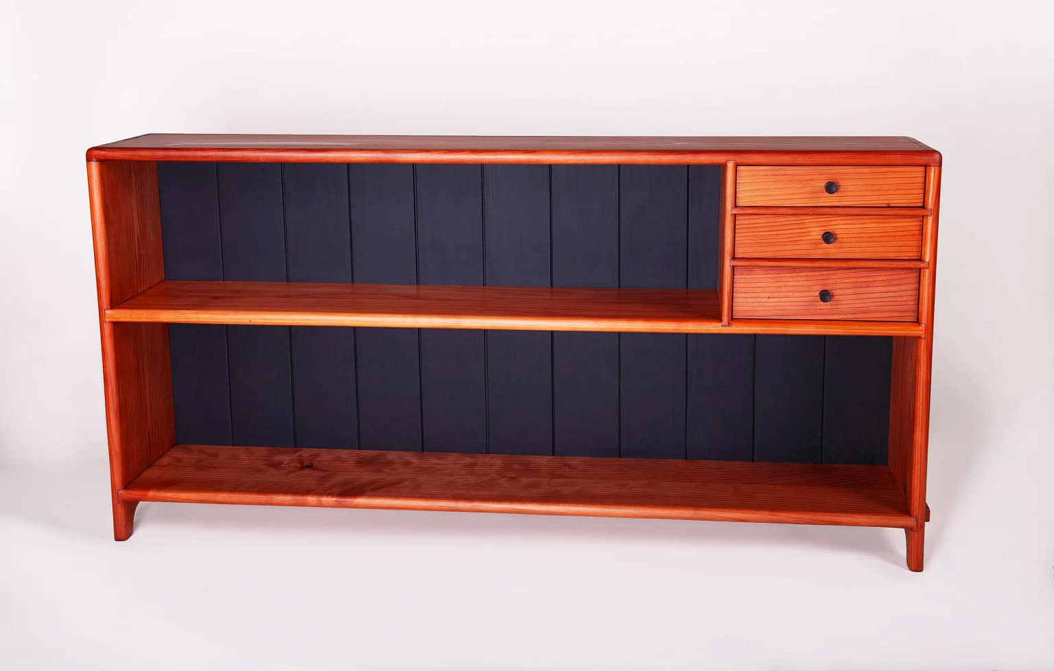 A low profile, free-standing redwood bookcase with two-shelves and three drawers. The middle shelf of the bookcase has 3 small drawers on the left hand side. The bookcase has a satin sheen and you can see that the back of the shelves behind the books is a solid black matte finish. It is a long bookcase with a low profile. It is 58" wide, 10 1/2 deep and 29" tall. this bookcase was milled, joined and crafted using only hand tools and traditional wood joinery. It has 5 coats of hand rubbed finish for a satin sheen. The piece was designed and crafted by Marc Springer a furniture maker in Woodland Hills, California.